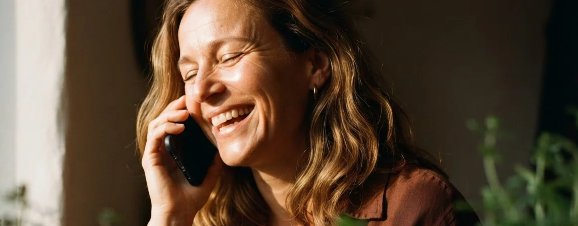 A woman laughing during a phone call at a sunlit window