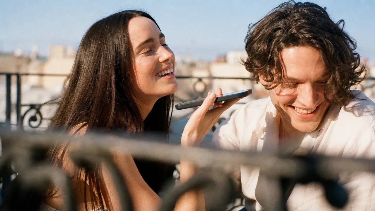 Two people leaning together over a phone on a rooftop terrace, laughing, city roofs behind them